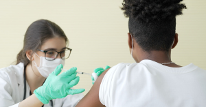 A seated pharmacist injects a vaccine into a seated patient's upper arm.