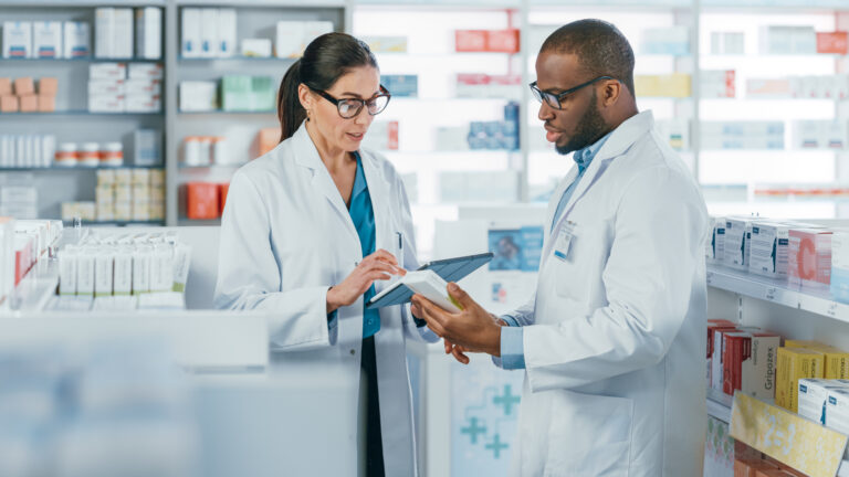Two pharmacists engaged in a discussion while standing in a pharmacy, with one pharmacist holding a tablet and both individuals focused on the screen. The pharmacy environment includes shelves of medication in the background, conveying a scene of professional collaboration and communication.
