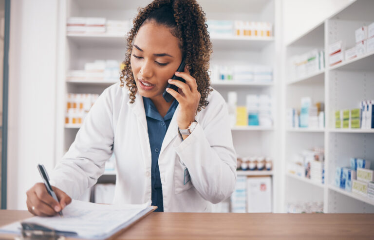 A pharmacist in a white coat speaks on the phone while writing on a patient information form at the pharmacy counter, with medication shelves visible in the background.