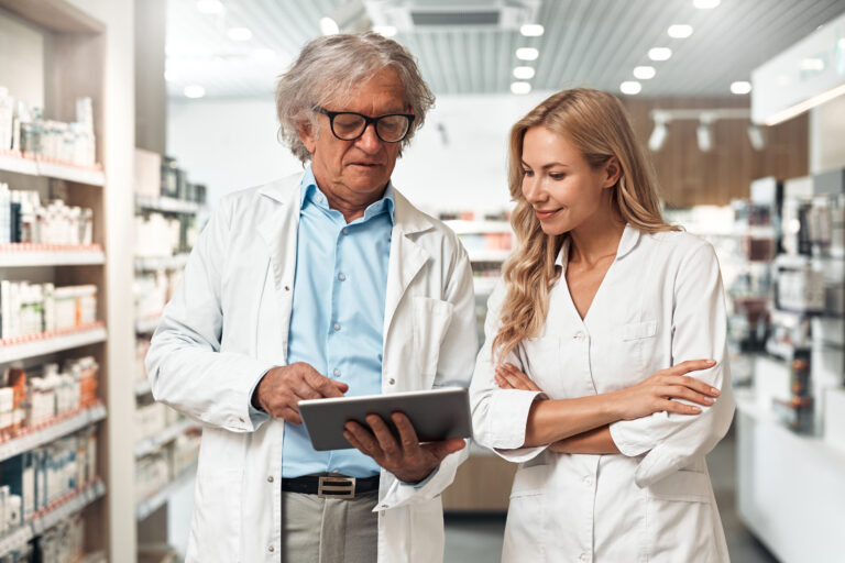 Male pharmacist showing information that can enhance the safety culture of the pharmacy on a tablet to female pharmacist. They both appear in a pharmacy setting.
